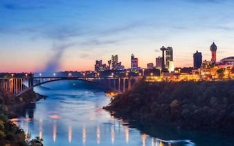 Niagara Falls USA skyline at dusk with illuminated cityscape and bridge over water.