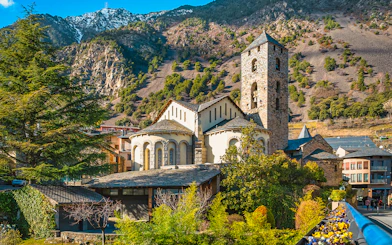 St. Esteve Church with Pyrenees Mountains in Andorra la Vella, surrounded by green trees.