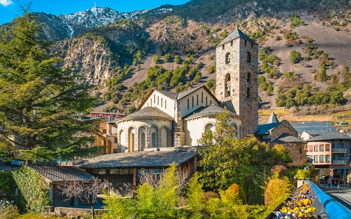 St. Esteve Church with Pyrenees Mountains in Andorra la Vella, surrounded by green trees.