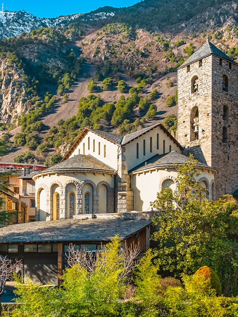 St. Esteve Church with Pyrenees Mountains in Andorra la Vella, surrounded by green trees.