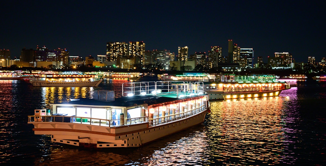 Houseboats with colorful lights on Tokyo Bay at night, city skyline in background.