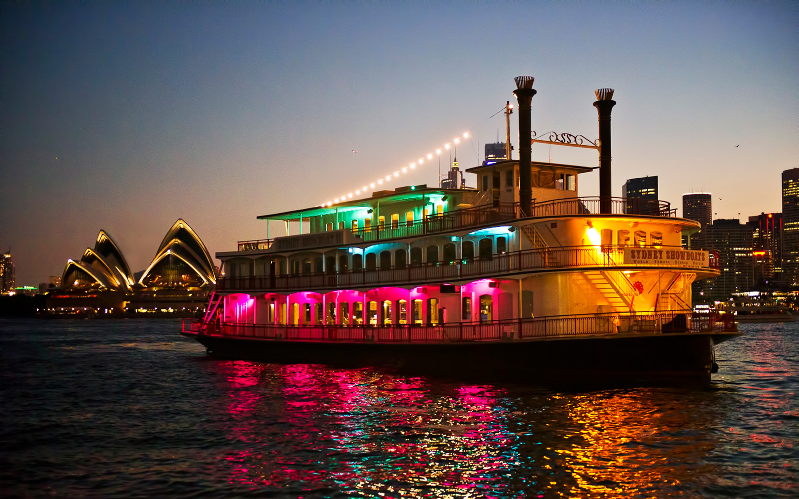 Cabaret cruise ship with colorful lights on Sydney Harbour near Opera House.