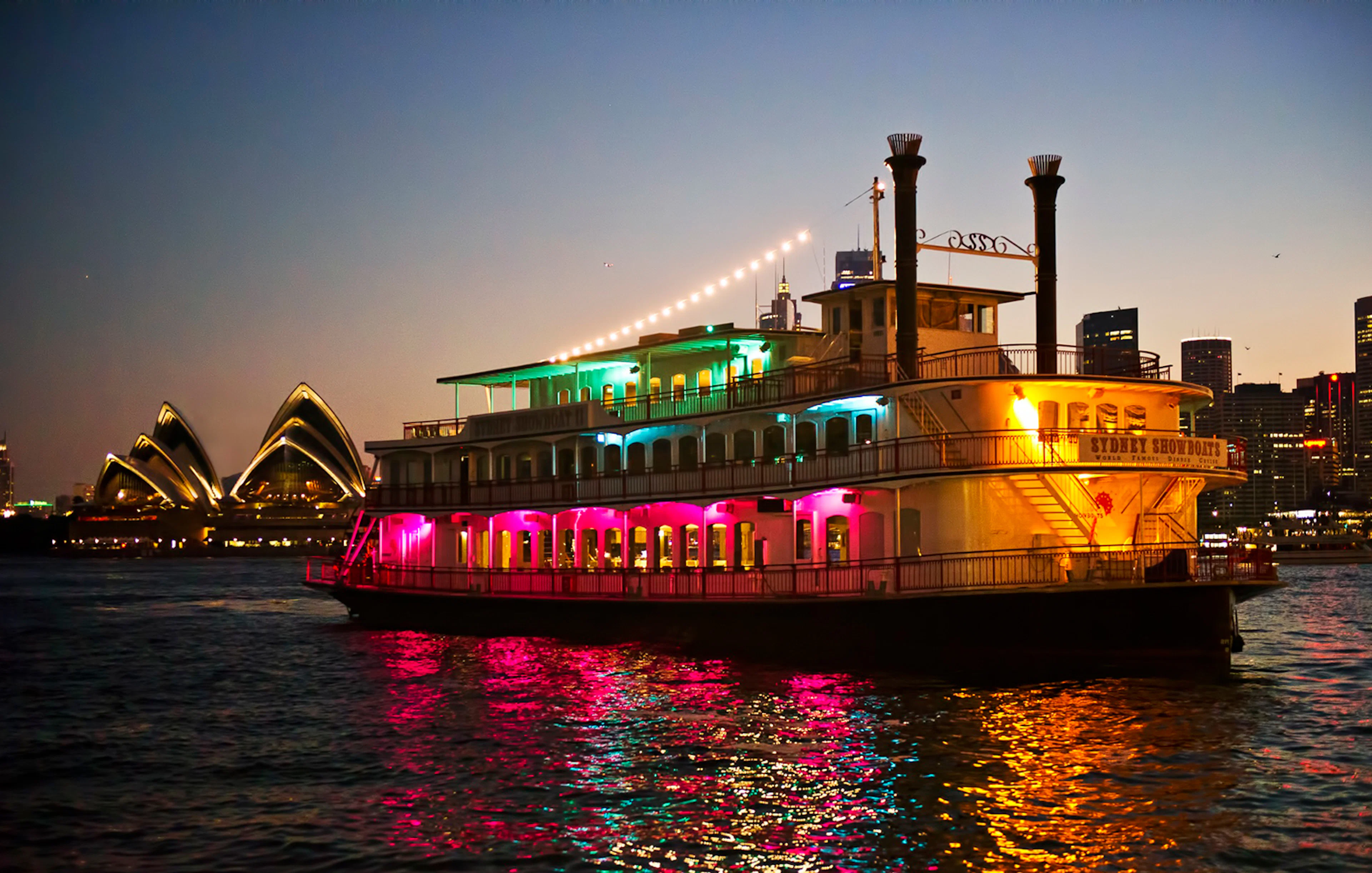 Cabaret cruise ship with colorful lights on Sydney Harbour near Opera House.