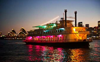 Cabaret cruise ship with colorful lights on Sydney Harbour near Opera House.