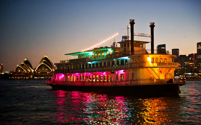 Cabaret cruise ship with colorful lights on Sydney Harbour near Opera House.