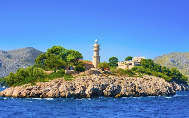 Lighthouse on rocky island with greenery, seen during catamaran trip in Alcudia.