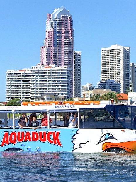 Aquaduck amphibious vehicle cruising on a river with city skyline in the background.