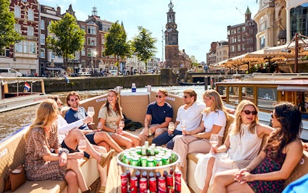 Group enjoying drinks on an Amsterdam canal cruise with historic buildings in the background.