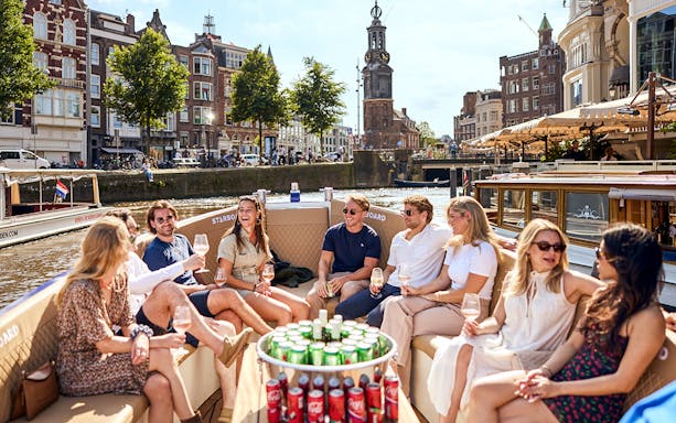 Group enjoying drinks on an Amsterdam canal cruise with historic buildings in the background.