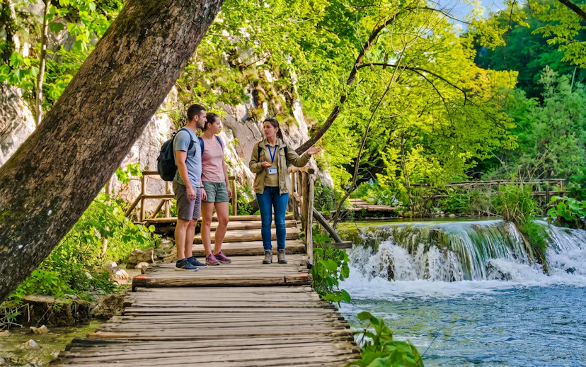 Tour guide explaining to tourists on a wooden path by waterfalls at Plitvice Lakes.