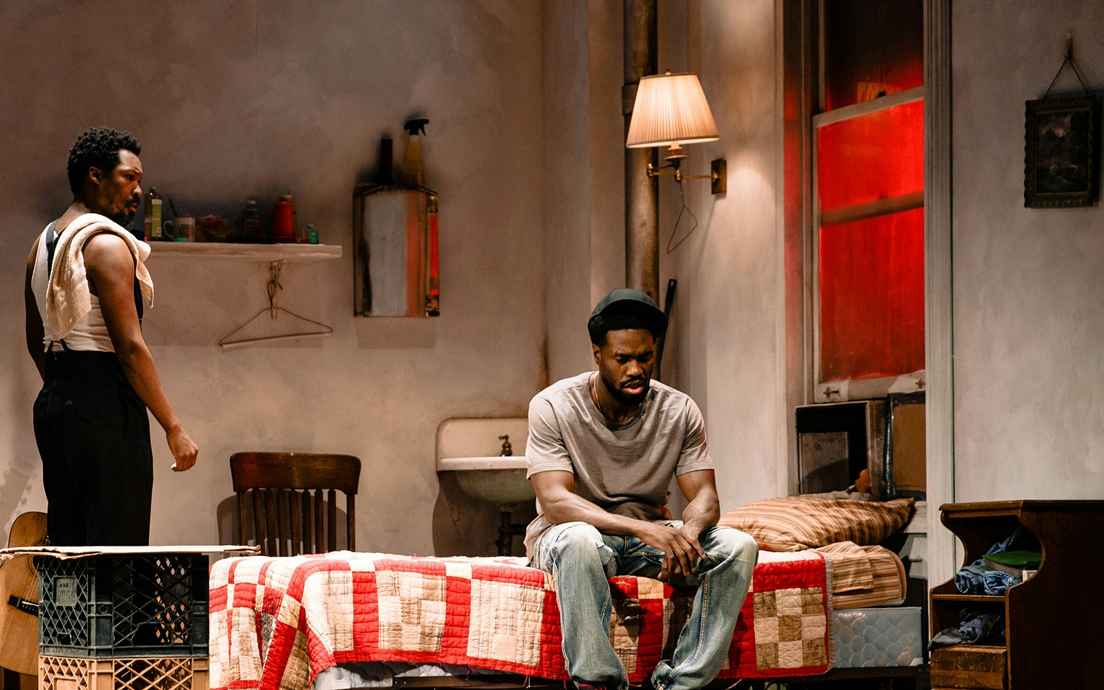 Two men in a dimly lit room, one sitting on a bed, the other standing, from the play Topdog/Underdog.