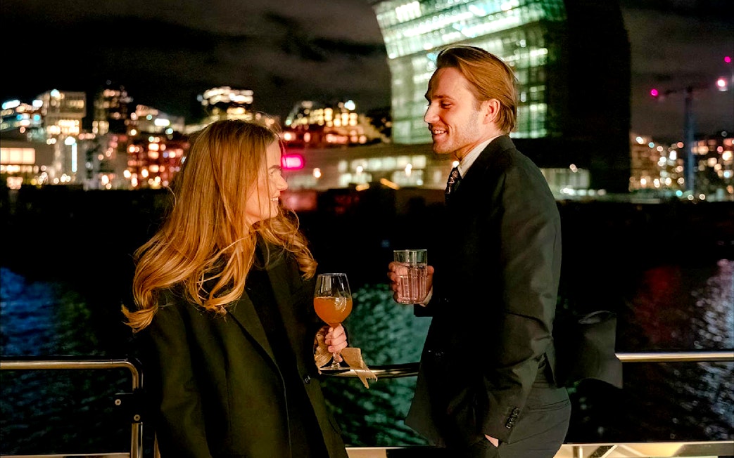 Couple enjoying drinks on an Oslo dinner cruise with city lights in the background.