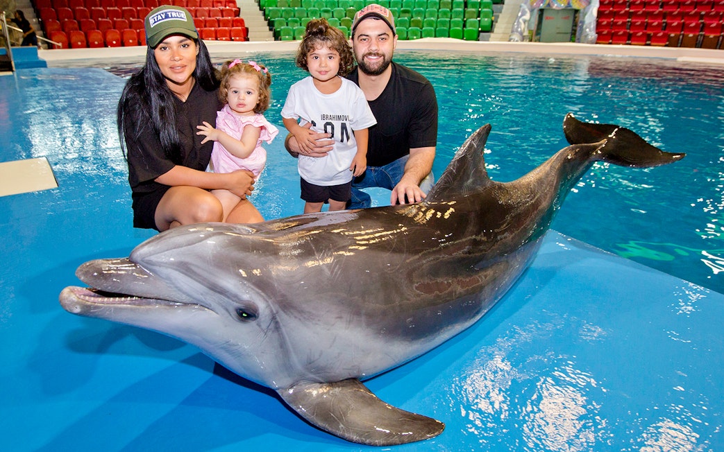 Tourists with children posing beside a dolphin at an aquarium.
