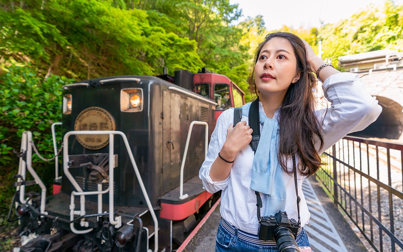 Woman standing beside Sagano train in lush forest setting.