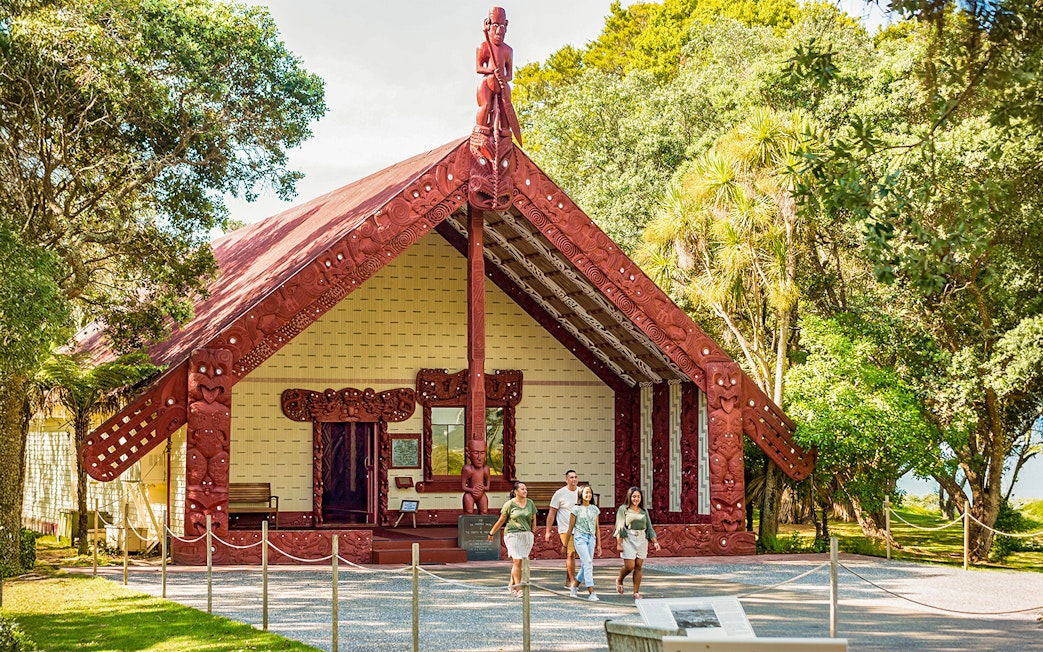 Te Whare Rūnanga meeting house with visitors walking in front, surrounded by lush greenery.