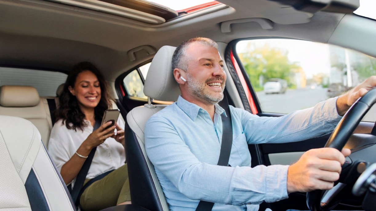 Driver and passenger smiling in a car, passenger using a smartphone.