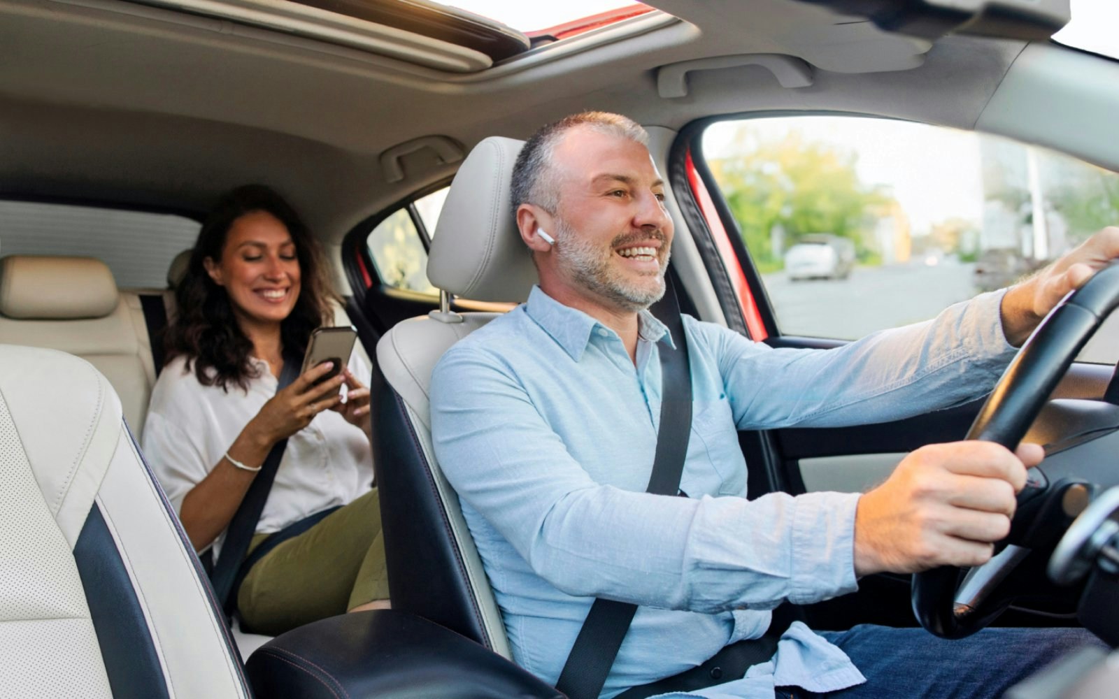 Driver and passenger smiling in a car, passenger using a smartphone.