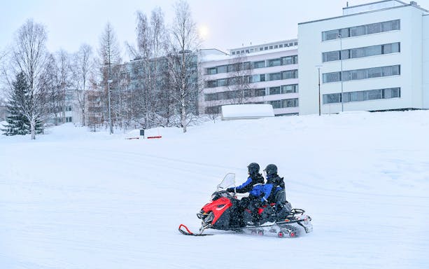 Snowmobile riders on a frozen lake in winter, Lapland, with snowy buildings in the background.