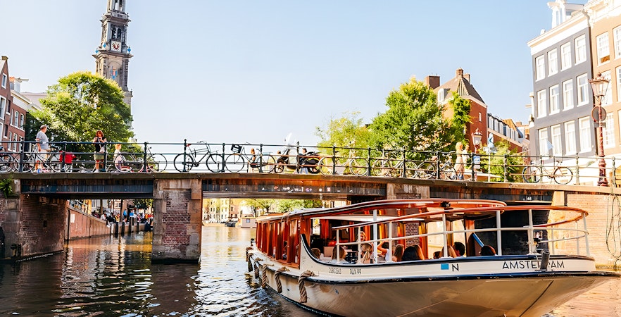 Saloon boat cruising under a bridge on an Amsterdam canal with Westerkerk tower in view.