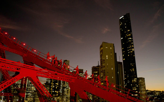 Climbers ascending Story Bridge in Brisbane at dusk with city skyline in background.
