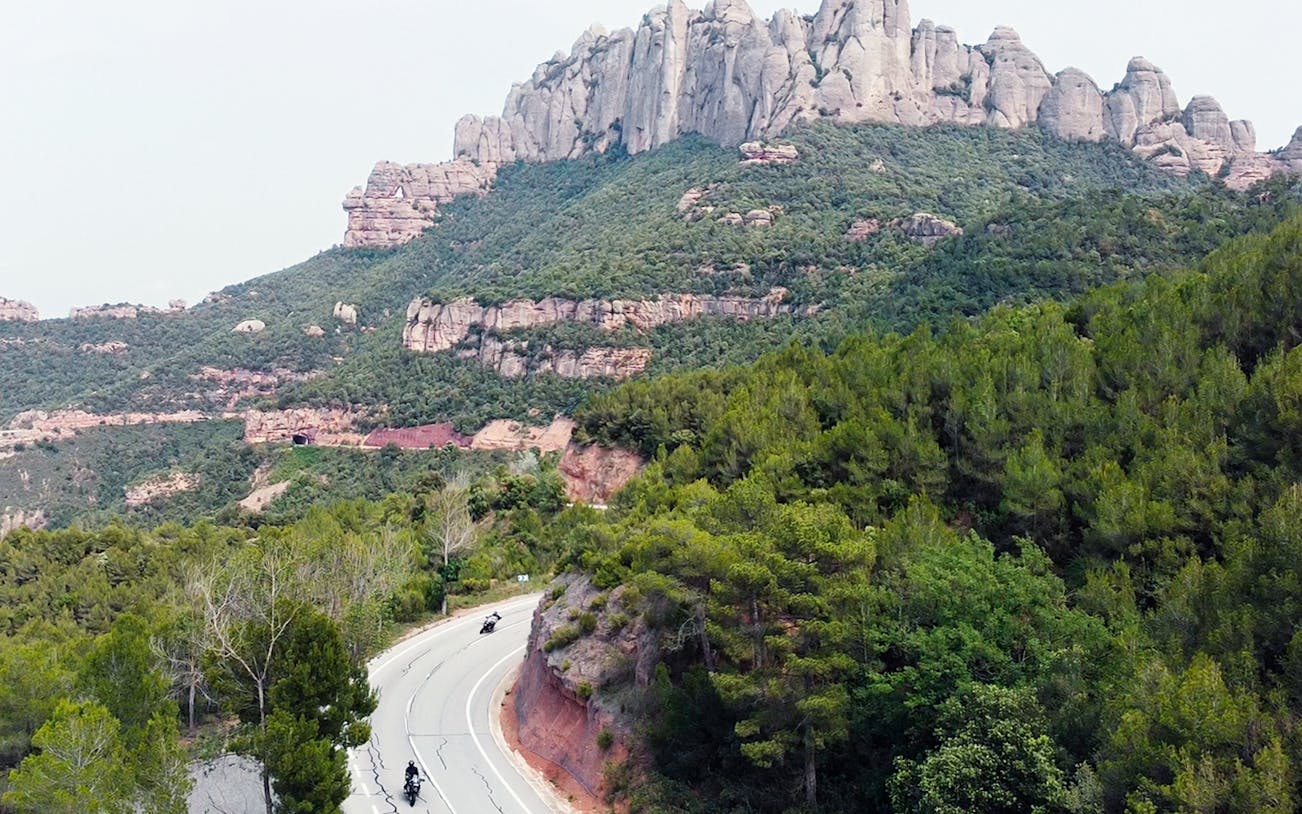 Motorcyclists riding on a winding road with Montserrat mountains in the background, near Barcelona.