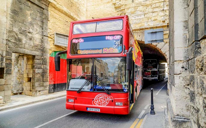 Red double-decker bus on Malta Hop-On Hop-Off tour passing through historic stone archway.