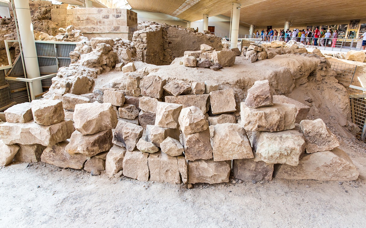 Ancient ruins at Akrotiri archaeological site, Santorini, with tourists observing.