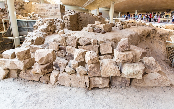 Ancient ruins at Akrotiri archaeological site, Santorini, with tourists observing.