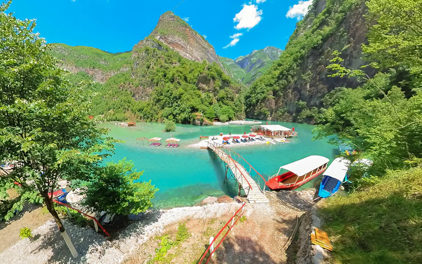 Komani Lake view with boats and dock on the Shala River, surrounded by lush mountains.