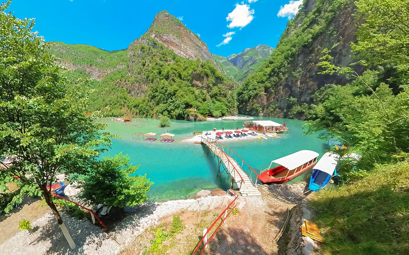 Komani Lake view with boats and dock on the Shala River, surrounded by lush mountains.