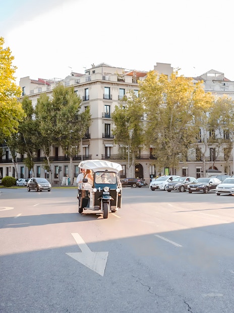 Electric tuk-tuk driving through a tree-lined street in Madrid.