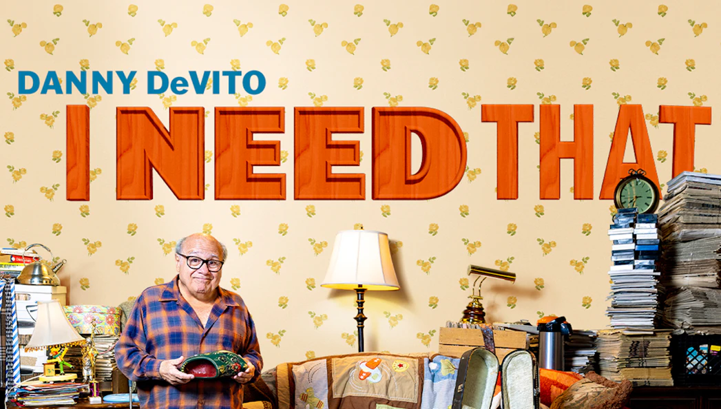 Man holding a mask in a cluttered room with "I Need That" text on the wall.