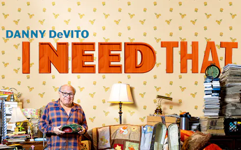 Man holding a mask in a cluttered room with "I Need That" text on the wall.