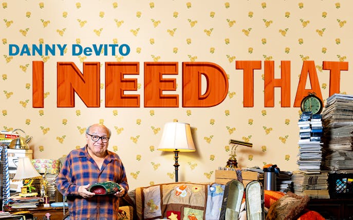 Man holding a mask in a cluttered room with "I Need That" text on the wall.