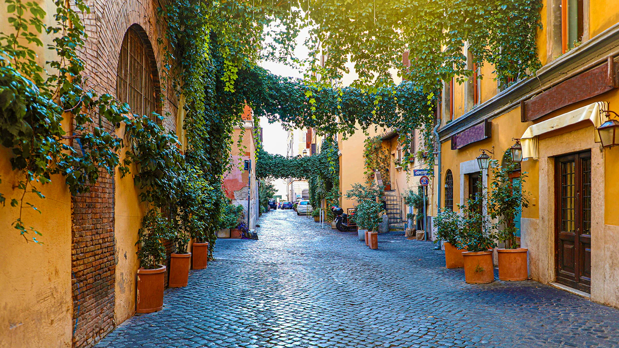 Cobblestone street in Trastevere, Rome, lined with ivy-covered buildings and potted plants.