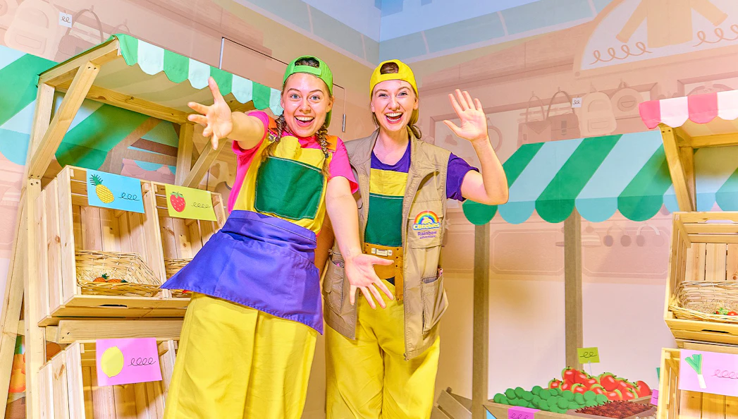 Two performers in colorful outfits at CBeebies Rainbow Adventure indoor market setup.