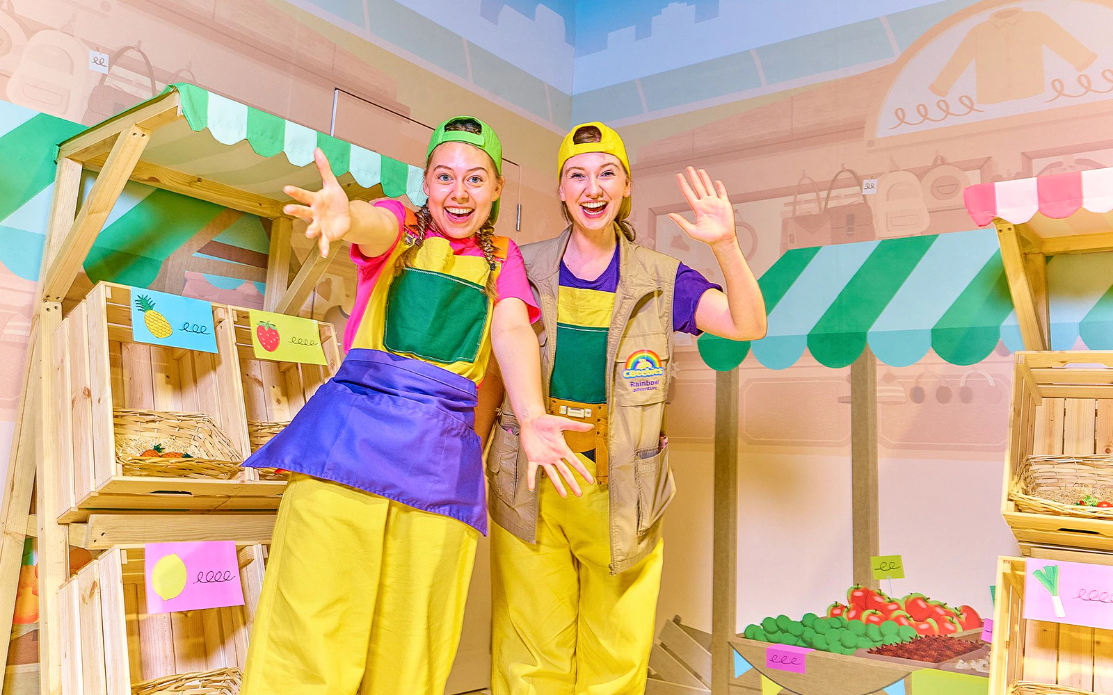 Two performers in colorful outfits at CBeebies Rainbow Adventure indoor market setup.