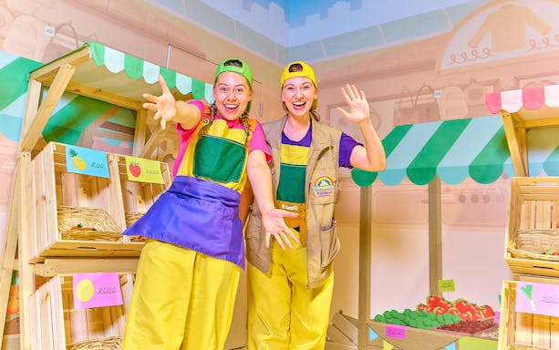 Two performers in colorful outfits at CBeebies Rainbow Adventure indoor market setup.