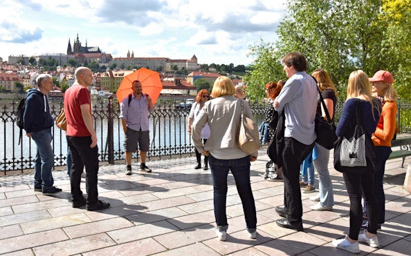 Tour group in Prague with guide near Vltava River, Prague Castle in background.
