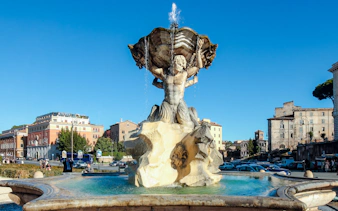 Fountain of the Tritons in Rome with surrounding buildings and clear sky.