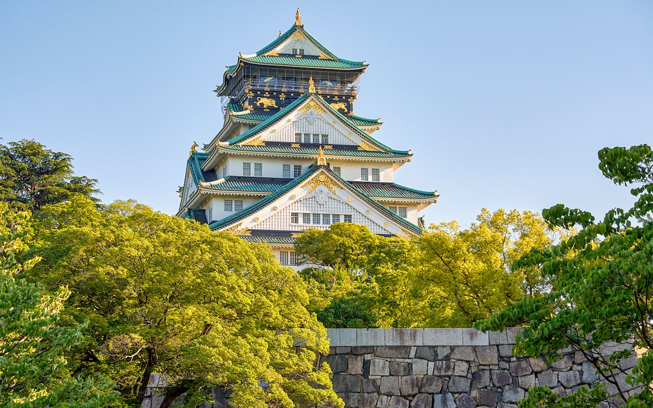 Osaka Castle exterior with lush green trees and blue sky.