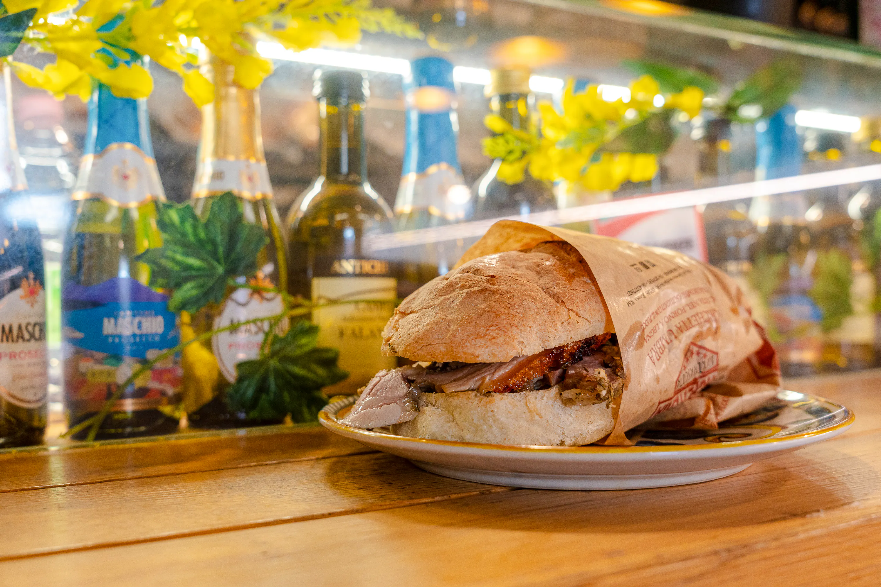 Panino con porchetta su piatto, bottiglie di vino sullo sfondo, Roma.