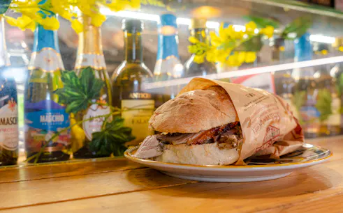 Panino con porchetta su piatto, bottiglie di vino sullo sfondo, Roma.