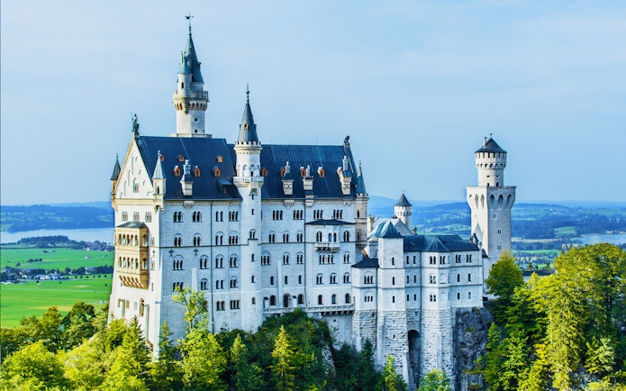 Neuschwanstein Castle in Bavaria, Germany, surrounded by lush greenery and distant hills.
