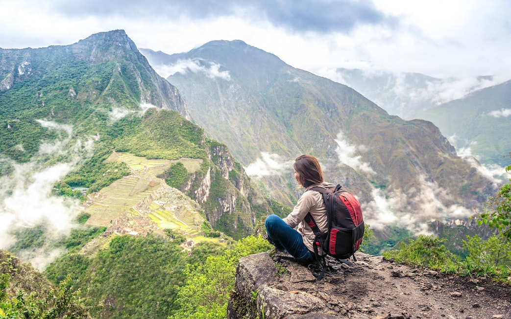 Person sitting on Wayna Picchu summit overlooking Machu Picchu ruins and surrounding mountains.