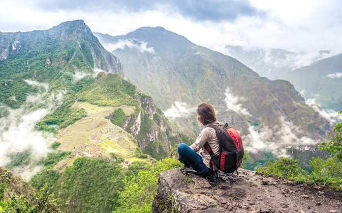 Person sitting on Wayna Picchu summit overlooking Machu Picchu ruins and surrounding mountains.