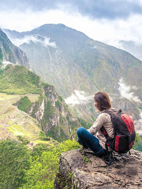 Person sitting on Wayna Picchu summit overlooking Machu Picchu ruins and surrounding mountains.