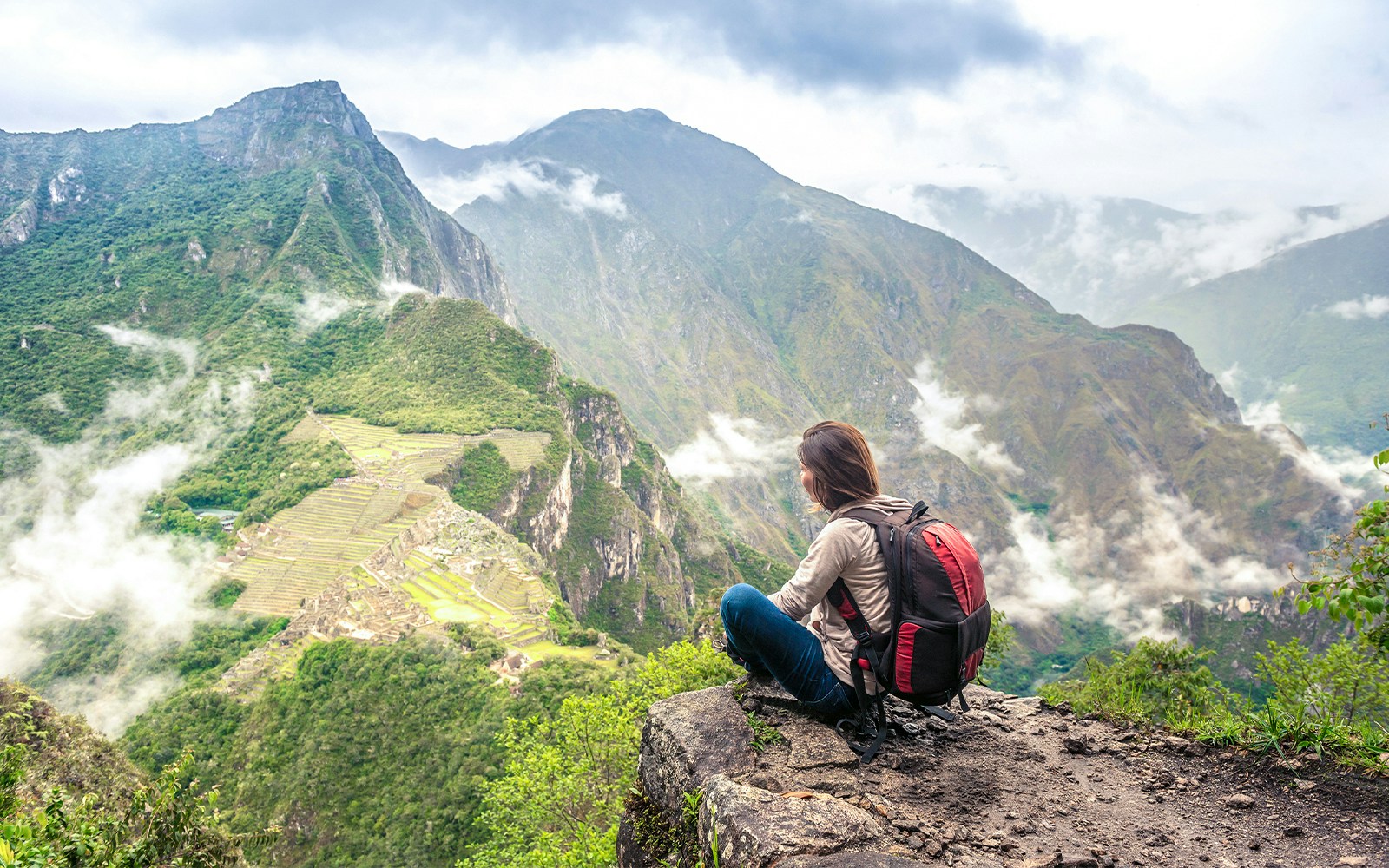 Person sitting on Wayna Picchu summit overlooking Machu Picchu ruins and surrounding mountains.