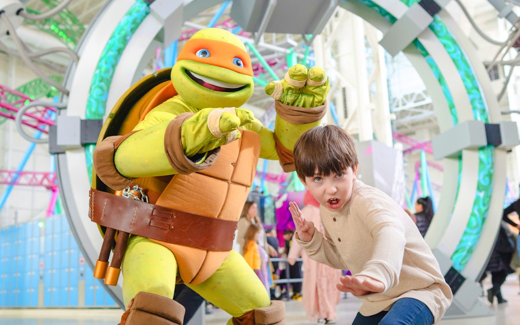 Child posing with Teenage Mutant Ninja Turtle character at an indoor amusement park.