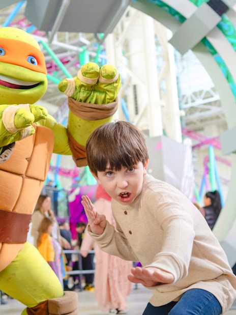 Child posing with Teenage Mutant Ninja Turtle character at an indoor amusement park.